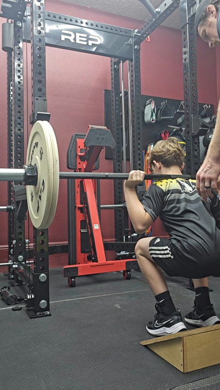 Young athlete back squatting with heels on a wedge and coach nearby